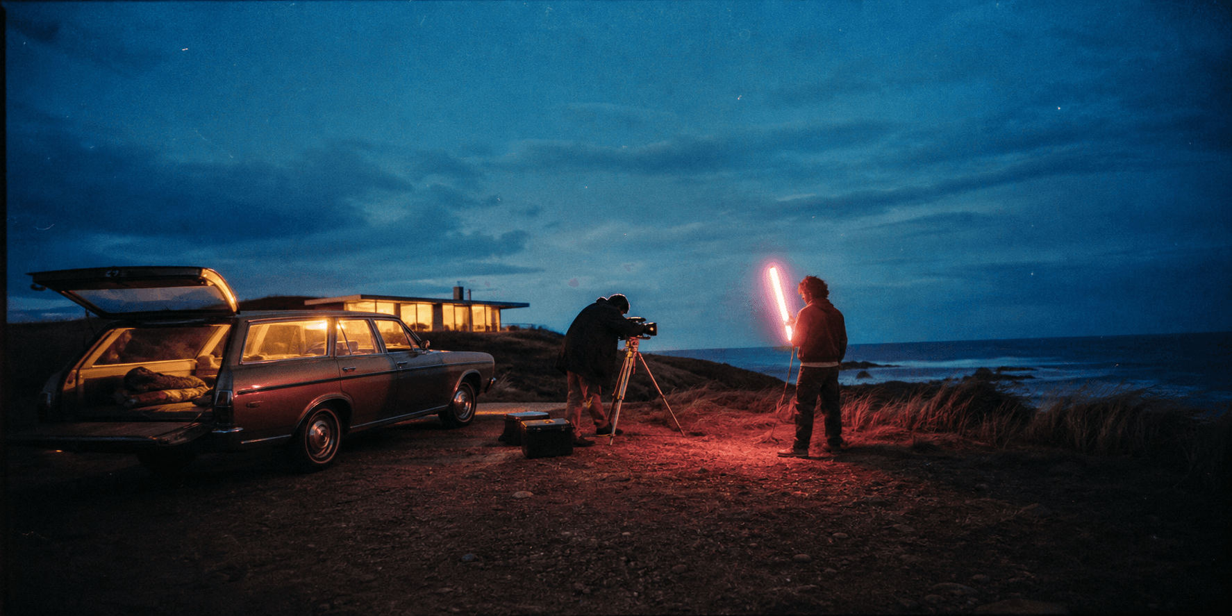 Grain-treated editorial image of a film crew beside a vintage station wagon on the coast at blue hour